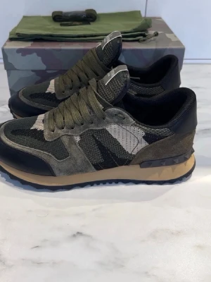 Valentino Garavani Rockrunner sneakers - Säljer ett par Valentino Garavani Rockrunner sneakers i camouflage med olivgröna🫒, beige och svarta toner. Skorna har snörning, detaljer i mocka och skinn samt en cool nubbad sula😎. Perfekt för dig som inte vill halka på vintern❄️