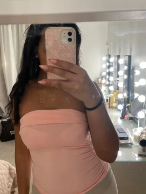 Ljusrosa bandeau tubtopp - Säljer en ljusrosa bandeau tubtopp med tight passform. Toppen är axelbandslös och har en enkel, stilren design som sitter snyggt mot kroppen. Perfekt att matcha med jeans eller kjol för en trendig look.