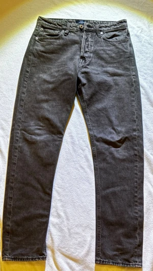 Bruna jeans från Jack & Jones - Säljer ett par bruna jeans från Jack & Jones med klassisk femficksdesign och knappgylf. Jeansen har raka ben och normal passform. Materialet är slitstarkt bomullstyg och färgen är en snygg mörkgrå ton. Perfekta till sneakers eller boots.