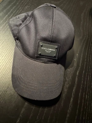 Dolce & Gabbana keps Äkta - Säljer en svart keps från Dolce & Gabbana med logopatch framtill. Klassisk böjd skärm och justerbar rem bak. Tillverkad i bomull med detaljer i skinn. Snygg och stilren accessoar som lyfter vilken outfit som helst.