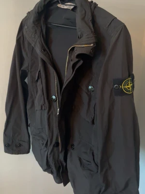 Svart parkajacka från Stone Island - Svart parkajacka från Stone Island med klassisk logga på ärmen. Jackan har flera fickor med knappar, dragkedja framtill och huva. Tillverkad i polyamid som ger en lätt och smidig känsla. Perfekt för dig som vill ha en stilren och funktionell jacka.
