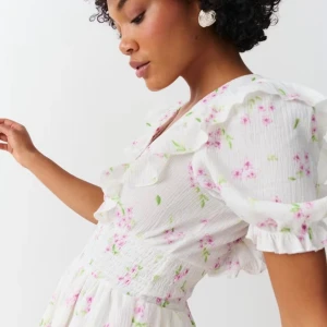 Vit blommig volangklänning med v-ringning - Supersöt vit kortklänning med rosa blommor och gröna blad, v-ringning och volangdetaljer längs axlar och kjol. Puffiga kortärmar med volangkant och smockad midja ger en romantisk vibe. Perfekt för sommarens alla tillfällen!