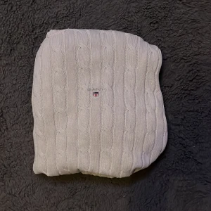 Vit kabelstickad tröja från Gant - Säljer en klassisk vit kabelstickad tröja från Gant med rund halsringning och diskret broderad logga på bröstet. Tröjan har långa ärmar och ribbade muddar för en snygg passform. Perfekt för lager-på-lager under höst och vinter.