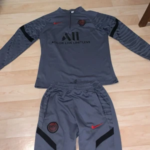 PSG långärmad träningsset Nike grå - Säljer ett grått långärmat träningsset från PSG med svarta och röda detaljer. Tröjan har halv dragkedja, PSG-logga och Accor Live Limitless-tryck framtill samt Visit Rwanda på ryggen. Ärmarna har subtilt zebramönster. Materialet är lätt och andas, perfekt för träning.