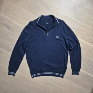 Marinblå half zip tröja från Hugo Boss - Snygg marinblå tröja från Hugo Boss med half zip och diskret logga på bröstet. Tröjan har ribbade muddar och krage med ljusgrå detaljer, samt vita ränder vid ärmslut och nederkant. Perfekt för dig som gillar stilren design och skön passform. Lite urtvättad runt dragkedjan annars i fint skick!