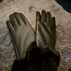 Svarta handskar med päls - Snygga svarta handskar med mjuk och fluffig pälsdetalj vid handleden. Handskarna har en stilren och klassisk design med slät yta och fem fingrar. Perfekta för kalla dagar när du vill hålla händerna varma och samtidigt se cool ut.