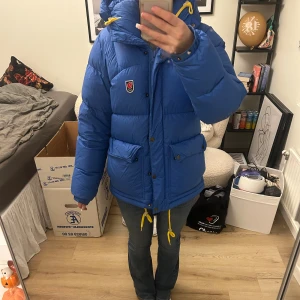 Blå dunjacka från Fjällräven - Blå dunjacka från Fjällräven. Jackan har stora fickor framtill, dragkedja och knappar. Klassisk Fjällräven-logga på bröstet och en riktigt puffig look. Perfekt för vintern! Säljer på grund av att jag söker en ny vinterjacka. Använd två vintrar men i bra skick! 