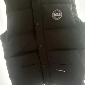 Svart dunväst från Canada Goose (black label) - Svart dunväst från Canada Goose med hög krage och tryckknappar framtill. Västen har två stora fickor med logotyp på ena fickan och ett broderat märke på bröstet. Perfekt för kalla dagar och har en stilren, quiltad design.