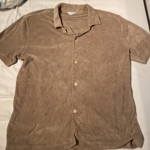 Beige frottéskjorta från East West - Säljer en beige skjorta från East West i mjuk frotté med korta ärmar och klassisk krage. Skjortan har knäppning framtill med fyra knappar och en avslappnad passform. Perfekt för dig som gillar chill och stilren vibe.