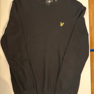 Svart stickad tröja från Lyle & Scott - Svart stickad tröja från Lyle & Scott med rund halsringning och den klassiska gula fågel-loggan  på bröstet. Tröjan har långa ärmar och ribbade muddar vid ärmslut och nederkant. 