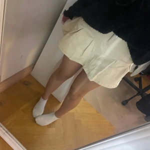 Beige kortkjol med hjärtdetaljer - Säljer en beige kortkjol med veckad design och fickor både fram och bak. Kjolen har dragkedja och knapp framtill samt söta hjärtformade detaljer på bakfickorna. Materialet är stadigt och känns som bomull, perfekt för dig som gillar en clean och söt look.