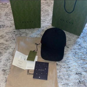 Svart Gucci keps med diskret logomönster - Snygg svart keps från Gucci med diskret GG-logomönster över hela kepsen och klassiska röda och gröna ränder på sidan. Justerbar rem baktill för perfekt passform. Materialet är bomull och polyester, vilket gör den både bekväm och stilren.