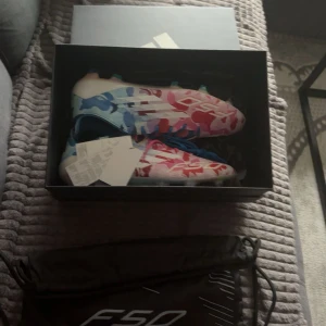 Adidas F50 färgglada fotbollsskor - Säljer ett par Adidas F50 fotbollsskor med unikt mönster i blått, rött och vitt. Skorna har snörning och är designade för att ge bra grepp på planen. Perfekta för dig som vill sticka ut med både stil och prestanda.