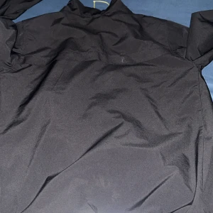 Svart vindjacka med dragkedja - Svart vindjacka med hel dragkedja framtill och elastiska muddar. Jackan har en slät yta och meshfoder på insidan för extra komfort. Perfekt för blåsiga dagar och enkel att matcha med olika outfits.