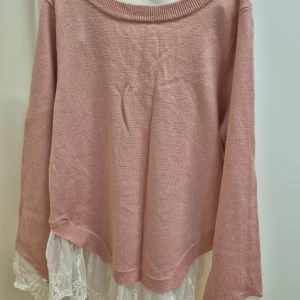 Rosa tröja med spets och rosett H&M XS - Supersöt rosa tröja med lång ärm, v-ringning i ryggen och stor vit rosett. Nedtill finns vit spetskant som ger en extra detalj. Tröjan är tillverkad i akryl och har en feminin vibe, perfekt för dig som gillar romantisk stil.