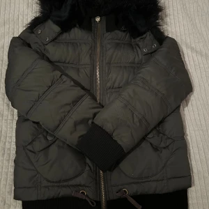 Svart pufferjacka från Big Star - Snygg svart pufferjacka från Big Star med huva och fuskpäls. Jackan har dragkedja framtill, ribbade muddar och två stora fickor med knapp. 