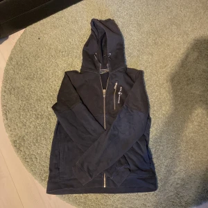 Svart och marinblå hoodie från Sail Racing med dragkedja - Svart hoodie från Sail Racing med hel dragkedja och broderad logga på bröstet. Tröjan har huva med snörning, två fickor fram och långa ärmar. Tillverkad i mjuk bomullsblandning, perfekt för kyliga dagar.