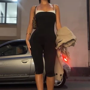 Capri byxdress med vita detaljer - Säljer en svart, figurnära byxdress med vita axelband och vit kant upptill. Byxdressen har trekvartsben och sitter tight över kroppen, vilket ger en snygg siluett. Väldigt strechigt material. Aldrig använt