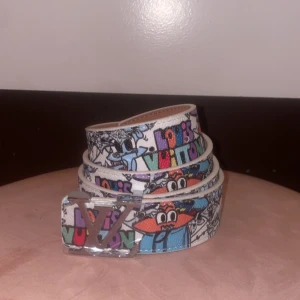Färgad bälte - Säljer ett unikt bälte med färgglada graffiti- och cartoonmotiv på vit botten. Bältet har en stor spänne i silver och coola detaljer i blått, orange, grönt och lila. Perfekt för dig som vill sticka ut med din stil!