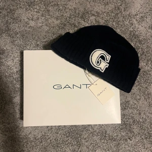 Mörk blå merinoull mössa från GANT - Mörk blå beanie från Gant med uppvikt kant och stor vit G-brodyr framtill. Klassisk och stilren design, perfekt för kalla dagar. Mössan är i mjukt material och har en tydlig chillkänsla.