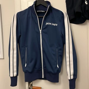 Palm Angels Zip - Snygg blå track jacket från Palm Angels med vita ränder längs ärmarna och vit dragkedja. Jackan har hög krage, två fickor med dragkedja och logga på bröstet. Perfekt för en sportig och trendig look.