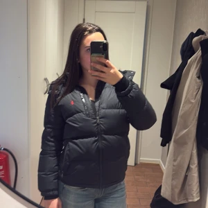 Svart pufferjacka från Polo Ralph Lauren - Säljer en svart pufferjacka från Polo Ralph Lauren med röd logga på bröstet. Jackan har dragkedja framtill, extra ficka med dragkedja på bröstet och hög krage. Perfekt för kalla dagar och riktigt snygg till jeans eller hoodies.