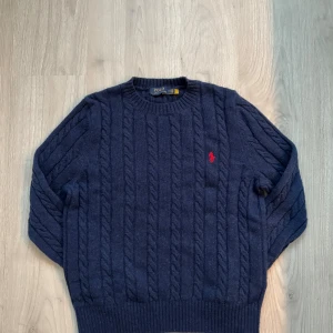 Mörkblå kabelstickad tröja Polo Ralph Lauren - Kabelstickad mörkblå tröja från Polo Ralph Lauren med klassisk röd broderad logga på bröstet. Tröjan har rund halsringning, ribbade muddar och är tillverkad i mjuk ull. Perfekt för dig som gillar en tidlös och stilren look. Köp nu och spara 115€