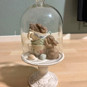 Dekorativ glasmonter med kaniner och tekoppar - Charmig dekorationsfigur med två söta kaniner, staplade tekoppar och dekorativa ägg under en glaskupa på en vacker fot. Perfekt som påsk- eller vårdekoration. Skicket är mycket gott, med endast små spår av slitage på foten. Ingen elektronik.