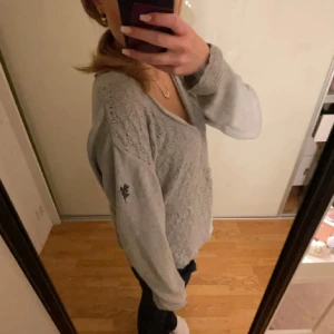 Ljusgrå stickad kofta med spetsmönster - Superfin ljusgrå stickad kofta med v-ringning och knappar framtill. Koftan har ett detaljerat spetsmönster och långa ärmar. broderad älva på ena ärmen.