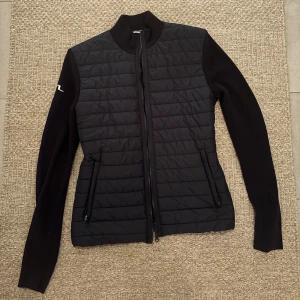 Svart jacka från J.Lindeberg S - Snygg mörkblå quiltad jacka från J.Lindeberg med ribbad krage och ärmar. Jackan har dragkedja framtill, två sidofickor med dragkedja och diskret logga på ena ärmen. Materialmix med stickade ärmar och quiltad front ger en modern look. Storlek S. Hör av dig vid frågor!