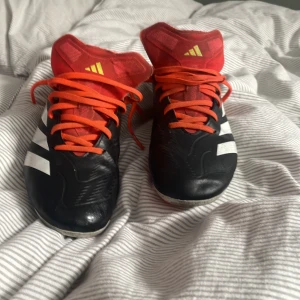 Adidas fotbollsskor svart/röd strl 38 - Säljer ett par Adidas fotbollsskor i svart med röda detaljer och orange snörning. Skorna har klassiska vita Adidas-ränder på sidorna och är tillverkade i syntetmaterial. Perfekta för dig som vill sticka ut på planen med snygg design och bra passform.