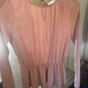 Rosa blus med spets och knyt i ryggen - Supersöt rosa blus med lång ärm, markerad midja och volang nedtill. Baksidan har djup v-ringning med spetsdetaljer och knytband i nacken. Perfekt för dig som gillar romantiska vibes och vill sticka ut lite extra.