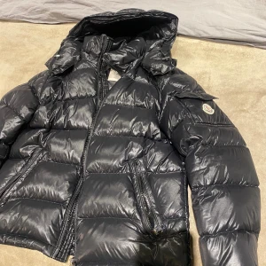 Svart Moncler Maya pufferjacka med huva - Säljer en svart pufferjacka från Moncler med glansig finish och huva. Jackan har dragkedja framtill, två snedställda fickor med dragkedja och klassisk Moncler-logga på ärmen. Perfekt för kalla dagar och riktigt snygg streetstil. KAN PRUTAS VID SNABB FÖRSÄLJNING!!!