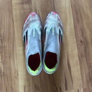 Adidas F50 fotbollsskor i vitt och rött - Säljer ett par Adidas F50 fotbollsskor i vitt med röda och svarta detaljer samt gula inslag. Skorna har en strömlinjeformad design utan klassisk snörning och är tillverkade i lätt syntetmaterial för maximal fart på planen. Har dock en liten skada i fram men det är inga större problem 