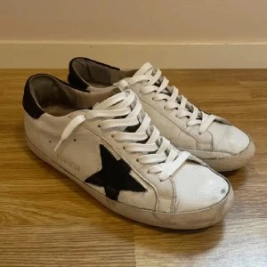 Golden Goose vita sneakers med svart stjärna - Golden Goose sneakers i vitt skinn med ikonisk svart stjärna på sidan och mörkbrun häl. Klassisk låg modell med vita snören och svart sula undertill. Snyggt slitna detaljer och retrovibe, perfekt för dig som gillar streetstyle. Storlek 42 tar byten 