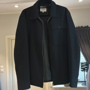 Svart jacka från Jack & Jones - Stilren svart overshirt från Jack & Jones i storlek M. Jackan har klassisk krage, bröstficka med knapp och dragkedja framtill. Materialet är mjukt och känns lite som ull, perfekt för lager på lager under höst och vinter. Enkel och clean look som funkar till det mesta.