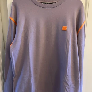 Lila långärmad tröja från Acne Studios - Säljer en oversized lila långärmad tröja från Acne Studios med orange logopatch på bröstet och orange detaljer vid axlar och nederkant. Ena ärmen har en grön quiltad panel som ger en unik look. Perfekt för dig som gillar streetwear och coola detaljer.