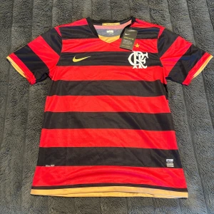 Flamengo Fotbollströja Nike  - Säljer en officiell Flamengo fotbollströja från Nike i storlek M. Tröjan har breda röda och svarta ränder, klubbmärke och Nike-logga på bröstet samt gulddetaljer vid ärmslut och hals. Tillverkad i lätt och ventilerande polyester med Nike FITDRY-teknologi.
