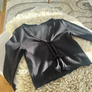 Kofta med knyt framtill - Svart topp från Lindex med knyt framtill och lång ärm. Toppen har en djup v-ringning och är tillverkad i ett mjukt, stretchigt material som sitter snyggt. Lite nopprig men annars i bra skick.