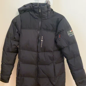 Svart pufferjacka från Sail Racing - Säljer en svart pufferjacka från Sail Racing med huva och dragkedja framtill. Jackan har logga på bröstet och ärmen, samt en liten ficka med röd dragkedja på ärmen. Perfekt för kalla dagar och riktigt snygg streetstil.