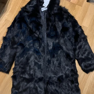 Svart pälsjacka från NA-KD - Snygg svart pälsjacka från NA-KD i storlek 34. Jackan har en fluffig och lyxig känsla med mjukt fuskpälsmaterial och klassisk krage. Perfekt för att lyfta din outfit under kyliga dagar. Stängs framtill och har en stilren look.