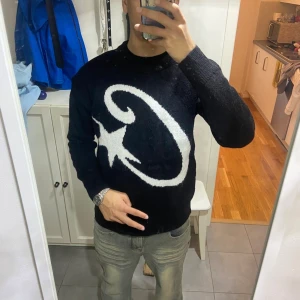 Cortiez  - Svart stickad tröja med en stor vit stjärna och böjd linje på bröstet. Tröjan har rund halsringning och långa ärmar. Perfekt för dig som vill sticka ut med en cool och unik design.