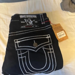 Svarta True Religion jeans med kontrastsömmar - Svarta jeans från True Religion med tydliga vita kontrastsömmar och stora fickor bak med lock och knapp. Klassisk logga på bakfickan och patch i midjan. Raka ben och cool streetstil, tillverkade i USA. Kan gå ner i pris vid snabb affär