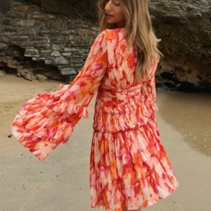 Nakd klänning  - Säljer en kort omlottklänning med v-ringning och långa volangärmar. Klänningen har ett färgstarkt mönster i rött, orange och rosa toner, med smockad midja och flera lager volanger. Materialet är lätt och luftigt, perfekt för varmare dagar. Storlek 36. Knappt använd