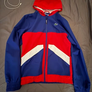 Blå och röd vindjacka från Lacoste - Snygg vindjacka från Lacoste i blått, rött och vitt med coola grafiska detaljer framtill. Jackan har huva, dragkedja och elastiska muddar. Perfekt för dig som gillar sportig stil och vill sticka ut med färg. Ikoniska Lacoste-loggan på bröstet.