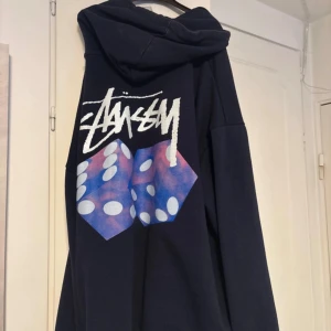 Stüssy marinblå hoodie med tärningsprint - Marinblå hoodie från Stüssy i 100% bomull med stor vit logga och färgglada tärningar på ryggen. Klassisk känguruficka framtill, huva med snörning och tryckt Stüssy-logga på bröstet. Perfekt för dig som gillar streetwear och vill sticka ut. Storlek L men har krympt till runt en M/S. Kan gå ner i pris vid snabb affär👍