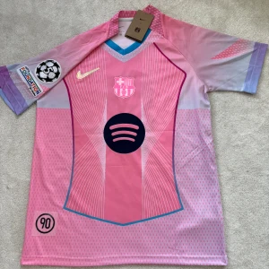 FC Barcelona rosa matchtröja Nike Size M - Säljer en FC Barcelona fotbollströja från Nike i rosa och lila toner. Tröjan har korta ärmar, V-ringning med blå detalj, tryckt klubbmärke, Champions League-märke och Spotify-logga. Tillverkad i lätt och ventilerande polyester.