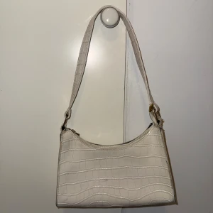 Vit axelväska med krokodilmönster - Snygg vit axelväska med krokodilmönstrad yta och guldfärgade detaljer. Väskan har en stilren form och stängs med dragkedja.