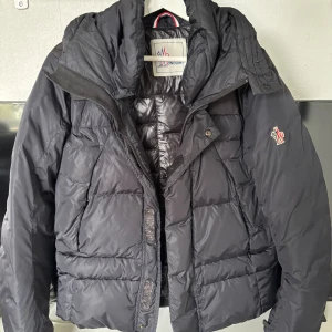  Moncler Grenoble jacka - Säljer en marinblå dunjacka från Moncler Grenoble med klassisk puffer-design. Jackan har huva, dragkedja och tryckknappar framtill samt Moncler-logga på ärmen. Jackan är vadderad för extra värme. Perfekt för kalla dagar. Den är köpt från kasperheat och använd 1 gång