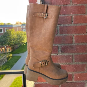 Bruna höga boots med klack och spänne - Säljer ett par snygga bruna höga boots med grov klack och plattformsula. Skorna har dekorativa spännen i metall och dragkedja på insidan för enkel påtagning. Materialet är mockaliknande och färgen är varm beige-brun. Perfekta för höst och vinter!